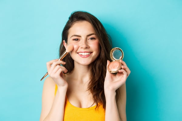 belleza-atractiva-mujer-joven-sonriendo-aplicando-maquillaje-pincel-mostrando-rubor-camara-pie-sobre-fondo-azul