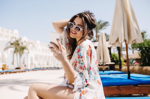 chica-morena-alegre-revisando-correo-redes-sociales-mientras-espera-sus-amigos-cerca-hotel-nadar-juntos-piscina-adorable-joven-gafas-sol-sentado-sillon-sosteniendo-telefono-blanco